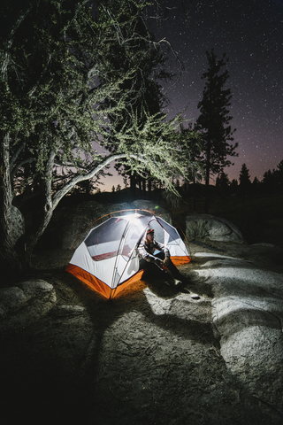 Hiker sitting in illuminated tent on rock by trees at night stock photo
