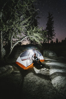 Wanderer, der nachts in einem beleuchteten Zelt auf einem Felsen bei Bäumen sitzt - CAVF28743