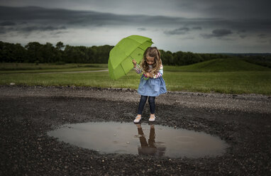 Mädchen hält Regenschirm, während sie in eine Pfütze auf der Straße gegen stürmische Wolken läuft - CAVF28746