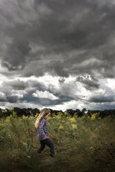 Unbekümmertes Mädchen, das inmitten von Pflanzen auf einem Feld gegen stürmische Wolken spazieren geht - CAVF28747