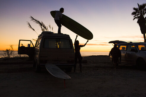 Silhouette eines Mannes, der einer Frau ein Surfbrett reicht, während er auf einem Minivan am San Onofre State Beach steht - CAVF28824