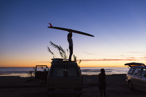Seitenansicht eines Mannes, der ein Surfbrett trägt, während er im Urlaub mit einer Frau am San Onofre State Beach auf einem Minivan steht - CAVF28825