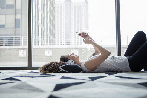 Junge Frau benutzt ihr Smartphone, während sie zu Hause auf dem Teppich am Fenster liegt - CAVF28887