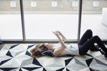 Young woman using smart phone while lying on carpet by window at home - CAVF28889