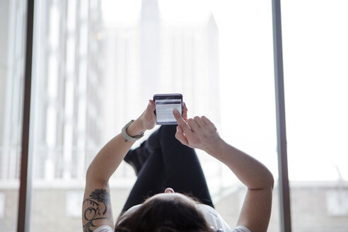 Woman using smart phone while lying against window at home - CAVF28891