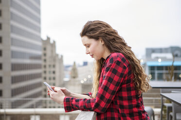 Seitenansicht einer Frau, die ihr Smartphone benutzt, während sie sich an das Geländer eines Terrassencafés lehnt - CAVF28896