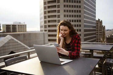Junge Frau benutzt ein Smartphone, während sie mit einem Laptop auf einem Tisch in einem Terrassencafé sitzt - CAVF28898