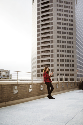 Frau in voller Länge, die ihr Smartphone benutzt, während sie sich an das Geländer der Terrasse eines Gebäudes lehnt, lizenzfreies Stockfoto