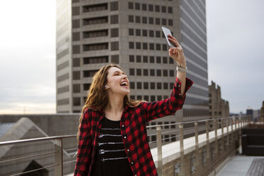 Cheerful young woman taking selfie while standing on terrace against building - CAVF28900