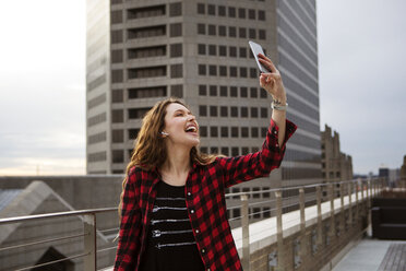 Fröhliche junge Frau, die ein Selfie macht, während sie auf einer Terrasse vor einem Gebäude steht - CAVF28900