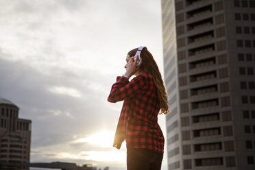 Side view of woman wearing headphones while standing against buildings - CAVF28902