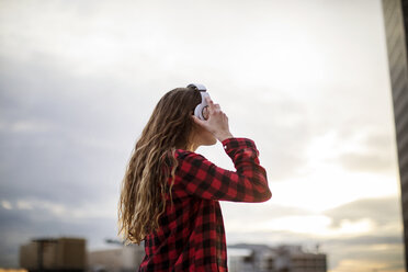 Side view of woman wearing headphones while standing against sky - CAVF28904