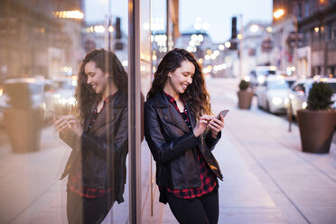 Smiling young woman using smart phone while standing on footpath by buildings - CAVF28909