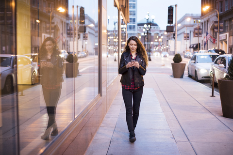 Junge Frau, die ihr Smartphone benutzt, während sie auf dem Fußweg bei Gebäuden spazieren geht, lizenzfreies Stockfoto