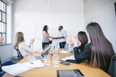 Businessman and businesswoman shaking hands while colleagues applauding in board room - CAVF29033