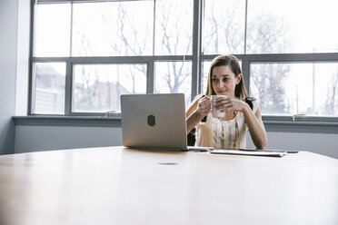 Geschäftsfrau, die ein Getränk in der Hand hält und auf einen Laptop-Computer schaut, während sie im Sitzungssaal sitzt - CAVF29060