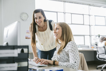 Lächelnde Geschäftsfrau, die neben einer Kollegin steht, die einen Computer am Schreibtisch im Büro benutzt - CAVF29066