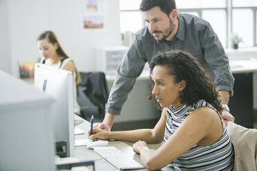 Businessman standing by female colleague using desktop computer at desk - CAVF29077