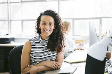 Portrait of smiling businesswoman sitting at desk with female colleague in background - CAVF29081