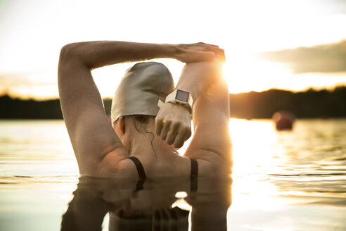 Rear view of female swimmer stretching arms in lake during sunset - CAVF29144