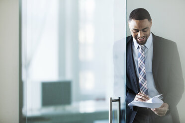 Happy businessman reading documents while standing by glass door in office - CAVF29227