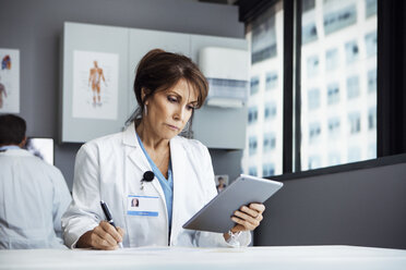 Female doctor using tablet computer while writing at desk in clinic - CAVF29291