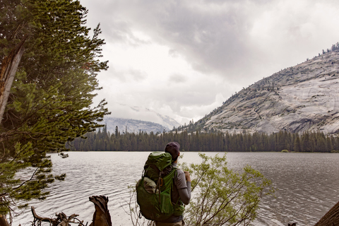 Wanderer mit Rucksack steht am Fluss im Yosemite-Nationalpark vor bewölktem Himmel, lizenzfreies Stockfoto