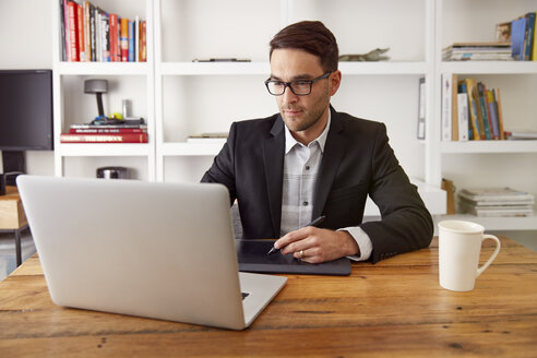 Geschäftsmann mit Grafiktablett und Laptop an einem Holztisch im Heimbüro - CAVF29383