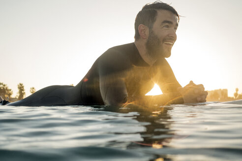 Glücklicher männlicher Surfer auf einem Surfbrett im Meer liegend bei Sonnenuntergang - CAVF29391