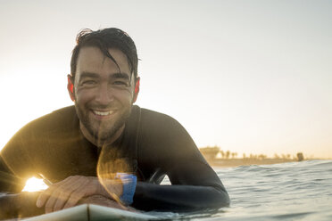 Porträt eines glücklichen männlichen Surfers, der bei Sonnenuntergang auf einem Surfbrett im Meer liegt - CAVF29392