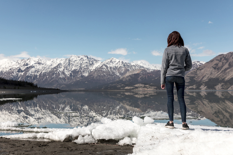 Full length of woman standing on snow by river while looking at view against mountains and sky stock photo