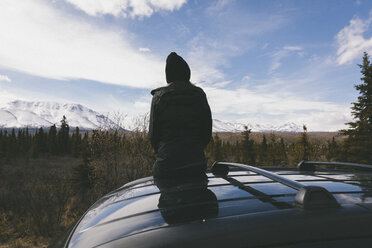 Rückansicht einer nachdenklichen, auf dem Autodach sitzenden Frau im Denali National Park vor schneebedeckten Bergen - CAVF29426