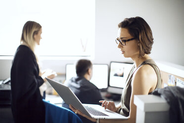 Geschäftsfrau mit Laptop, während Kollegen im Kreativbüro arbeiten - CAVF29500