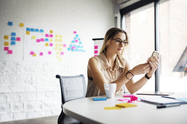 Thoughtful businesswoman using phone while sitting at table in creative office - CAVF29524