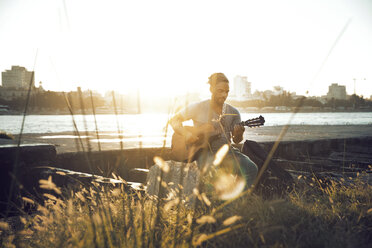 Mann spielt Gitarre, während er am Seeufer sitzt - CAVF29562