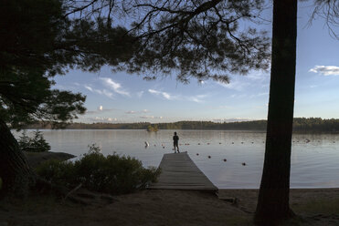 Silhouette boy standing on jetty over lake against blue sky - CAVF29590