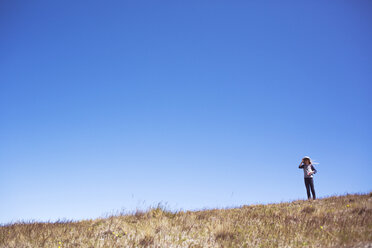 Girl looking through binoculars while standing on grassy field against clear blue sky - CAVF29610