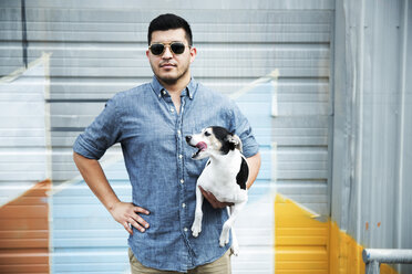 Mann mit Sonnenbrille, der einen Hund trägt, während er auf der Straße an der Wand steht - CAVF29628