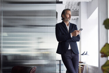 Thoughtful businessman looking away while holding smart phone against glass wall in office - CAVF29689