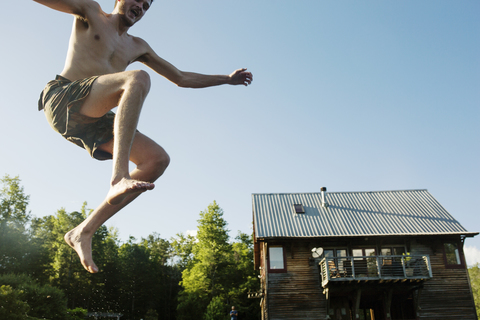 Niedriger Blickwinkel von hemdsärmeligen Mann springt in den See gegen Blockhaus und klaren Himmel, lizenzfreies Stockfoto