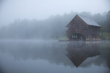 Symmetry view of log cabin by lake against trees during foggy weather - CAVF29730