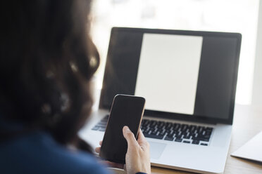 Abgeschnittenes Bild einer Geschäftsfrau, die ein Mobiltelefon benutzt, während sie an einem Laptop im Büro sitzt - CAVF29780