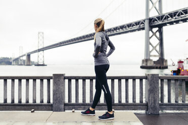 Seitenansicht einer Sportlerin mit Blick auf die Oakland Bay Bridge, während sie auf dem Fußweg steht - CAVF29809