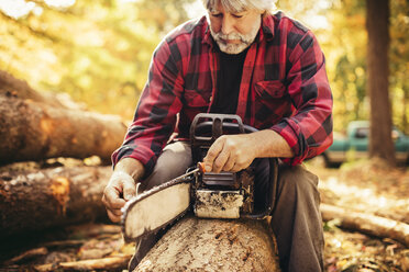 Mature male lumberjack examining chainsaw while sitting on log in forest - CAVF29863