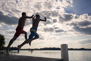 Father and son jumping from pier at lake against sky - CAVF29882