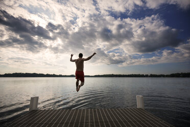 Rear view of man jumping into lake against cloudy sky - CAVF29886