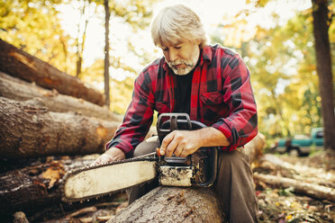 Male lumberjack examining chainsaw while sitting on log in forest - CAVF29944