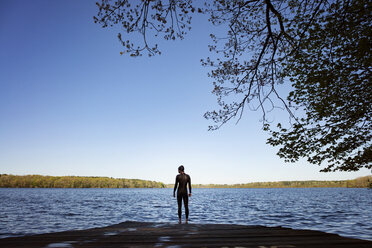 Rückansicht einer Schwimmerin, die auf der Promenade am See steht, gegen einen klaren blauen Himmel - CAVF29973