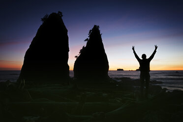 Silhouette eines Mannes, der bei Sonnenuntergang mit erhobenen Armen am Ufer steht - CAVF30016