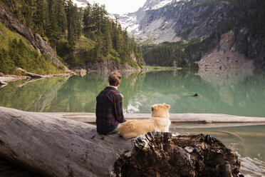Rückansicht eines Mannes und eines Hundes, die auf einem umgestürzten Baum am Blanca-See sitzen - CAVF30062